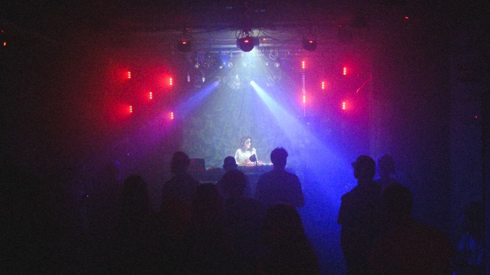 Koncert w klubie, na scenie dziewczyna za didżejską konsoletą. Jest ciemno, scenę oświetlają czerwone i niebieskie światła.