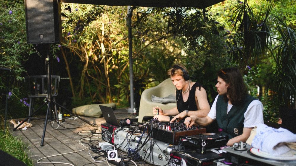 Dwaj młodzi mężczyźni siedzą na tarasie otoczonym bujną roślinnością, obsługują elektroniczne urządzenia muzyczne. Jeden z nich ma na głowie słuchawki.