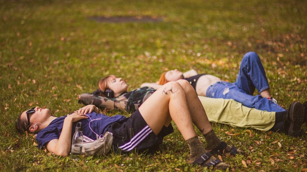 Trzy młode dziewczyny leżą na zielonej trawie słuchając muzyki. Mają zamknięte oczy, sprawiają wrażenie zrelaksowanych.