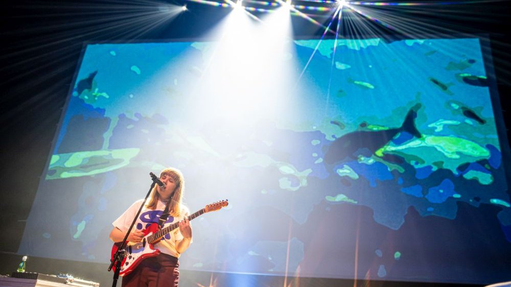 Iwona Skwarek śpiewa do mikrofonu grając na gitarze. Za jej plecami wizualizacje z morskim motywem.
