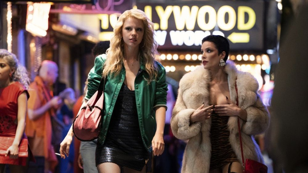 Dwie młode kobiety idą przez miasto oświetlone neonami. Jedna z nich pali papierosa, druga nosi białe futro i mocny makijaż.