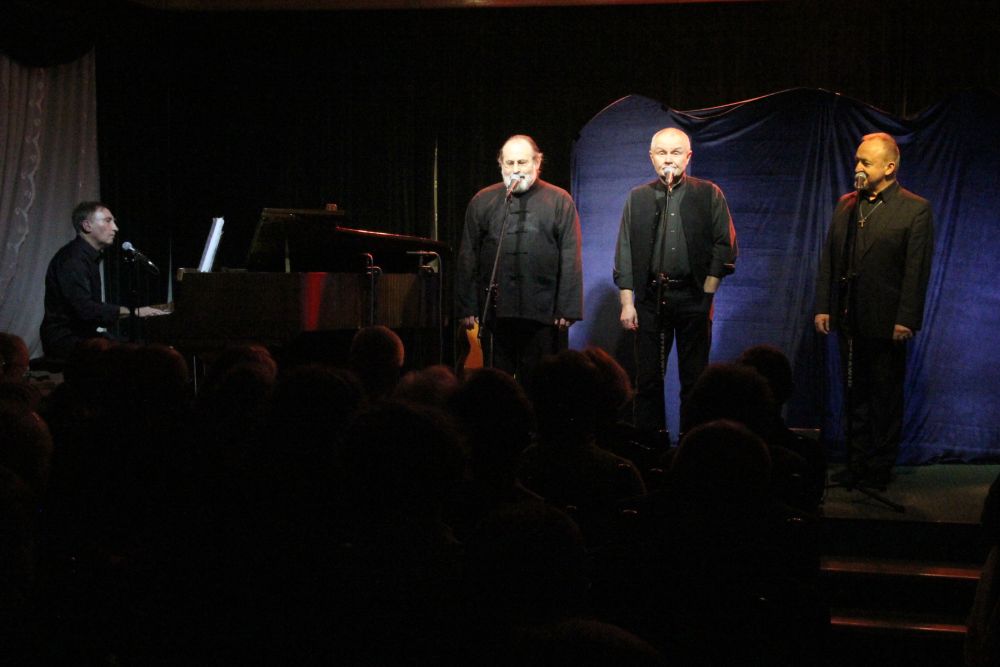 Czterech mężczyzn ubranych na czarno występuje na scenie. Trzech z nich śpiewa, jeden gra na fortepianie. - grafika artykułu