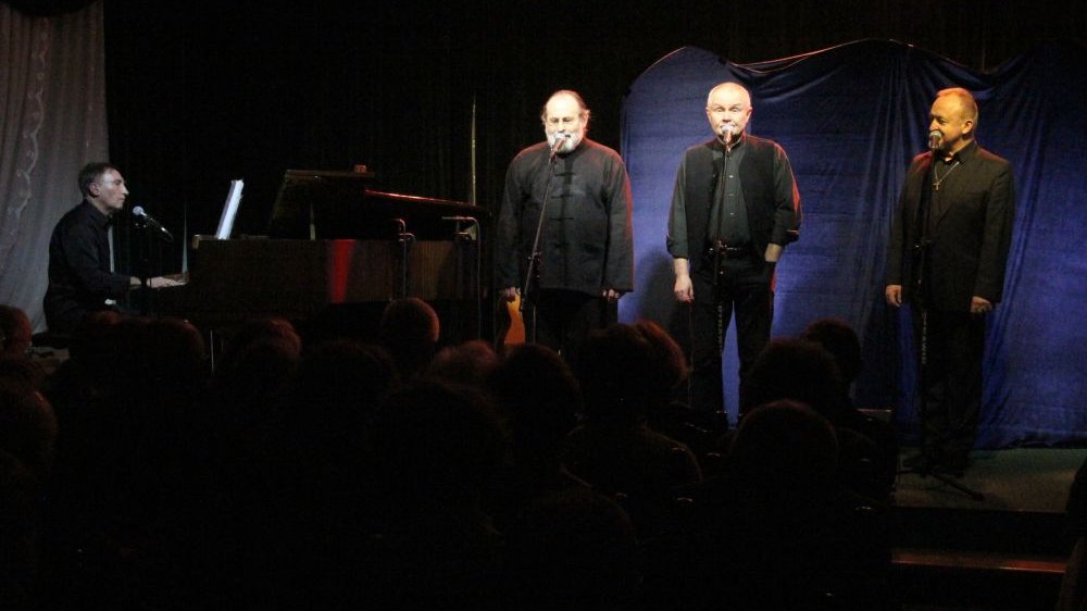 Czterech mężczyzn ubranych na czarno występuje na scenie. Trzech z nich śpiewa, jeden gra na fortepianie.