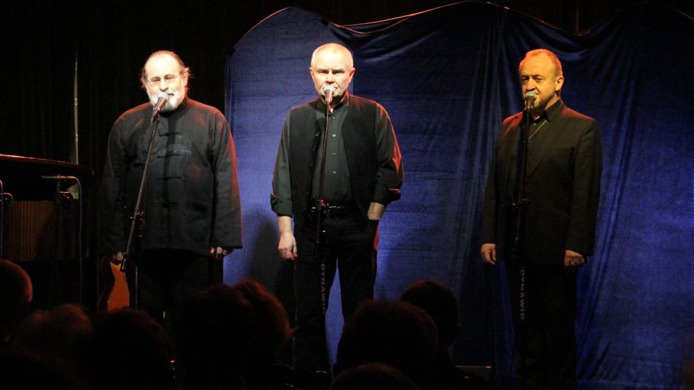 Trzech mężczyzn na scenie, śpiewają do mikrofonów.