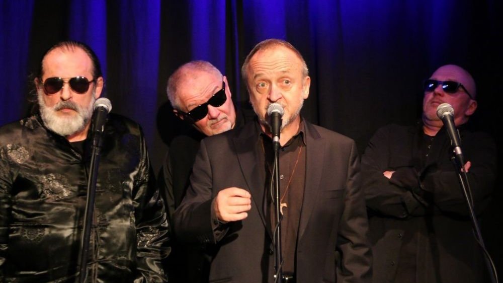 Czterech mężczyzn na scenie. Trzech z nich ma założone okulary przeciwsłoneczne.