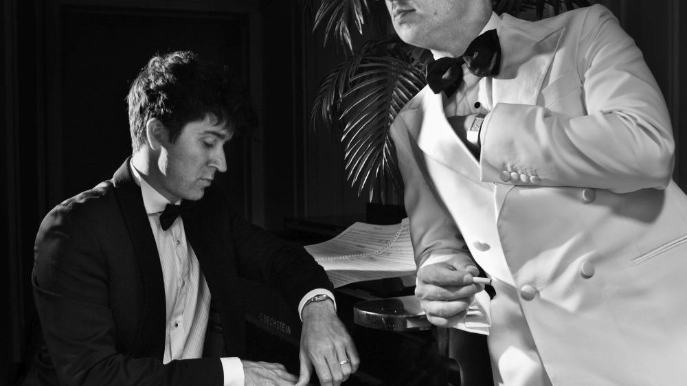 Dwóch elegancko ubranych mężczyzn przy fortepianie.