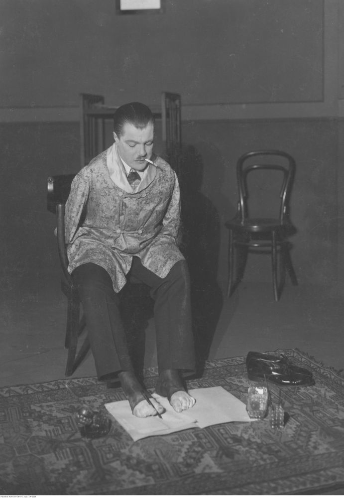 Mężczyzna bez rąk pisze na papierze leżącym na ziemi używając stóp. Siedzi na krześle, w ustach ma papierosa. - grafika artykułu