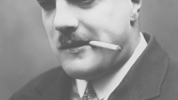 Elegancki mężczyzna z wąsem pali papierosa lewym kącikiem ust.
