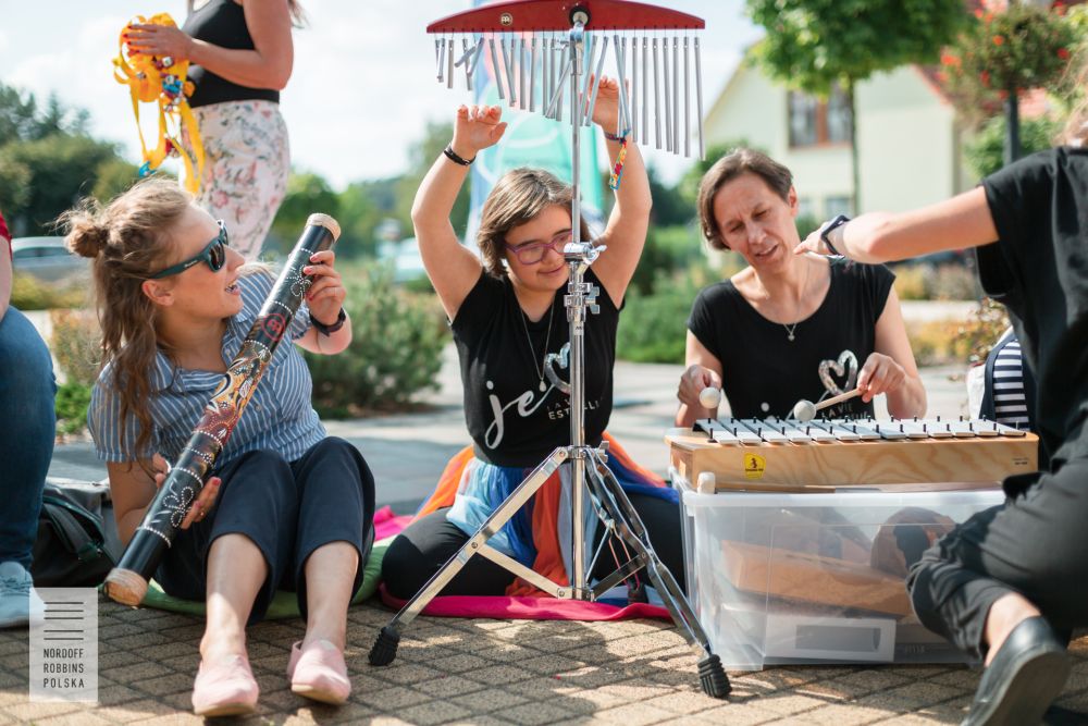 Kilka kobiet siedzi w słonecznym plenerze przy instrumentach, jedna z nich gra na cymbałkach, inna na dzwonkach, jeszcze inna trzyma tubę. - grafika artykułu