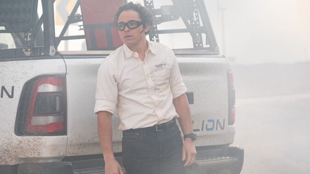 Mężczyzna w białej koszuli i okularach ochronnych stoi za samochodem. Wokół wiruje kurz i pył.