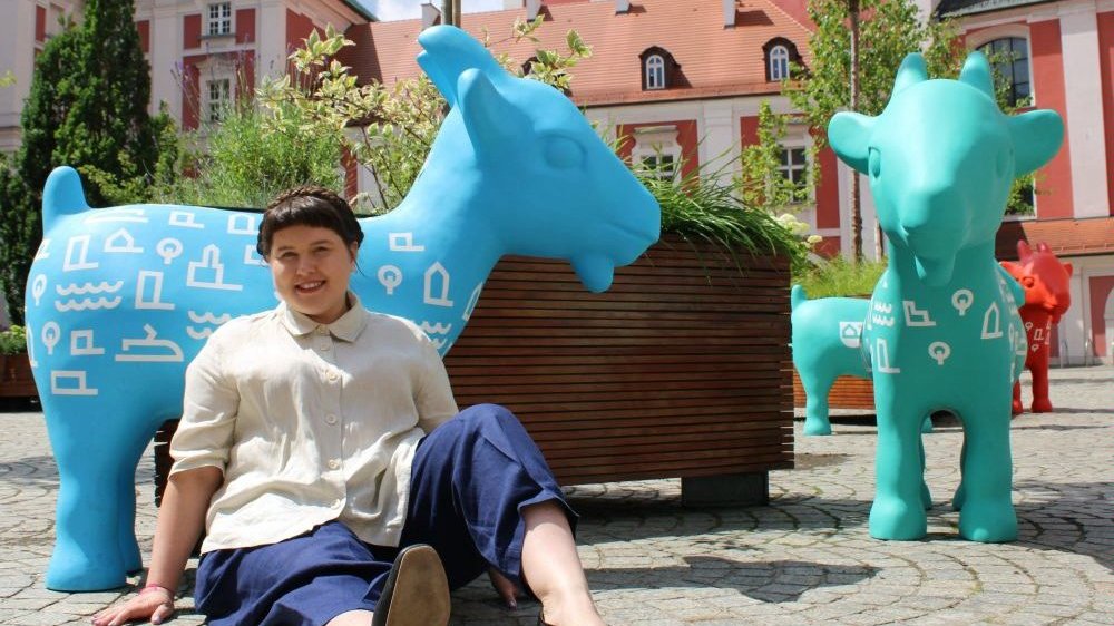 Uśmiechnięta dziewczyna siedzi na bruku przy kolorowych figurkach koziołków poznańskich na dziedzińcu Urzędu Miasta Poznania.