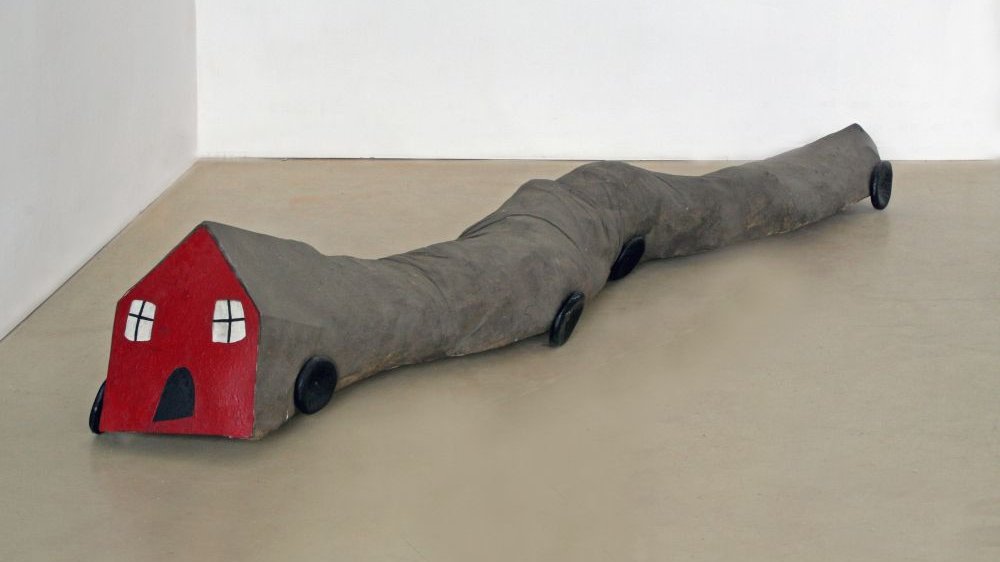 Przypominający węża na kółkach, z czerwonym domkiem zamiast głowy, pluszowy obiekt leży na podłodze muzeum.