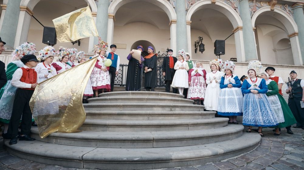 Ludzie w tradycyjnych strojach bamberskich stoją na schodach ratusza poznańskiego. Dwoje z nich trzyma złote flagi.