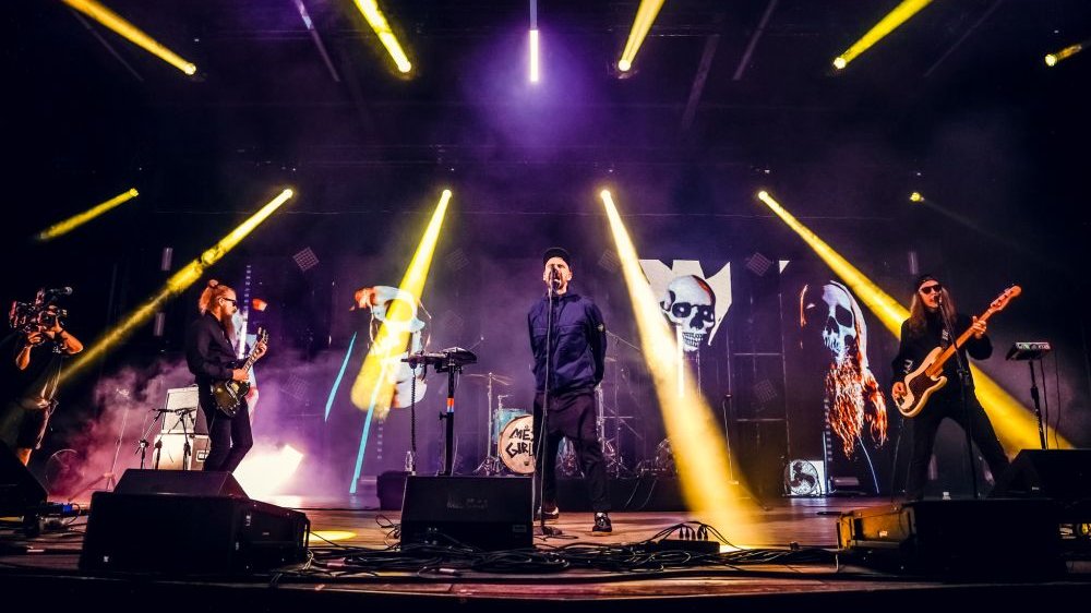 Muzycy na scenie. Zza ich pleców scenę oświetlają żółte smugi świateł.