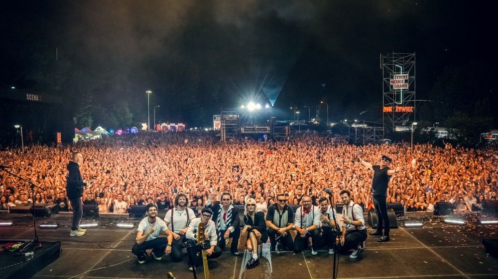 Zespół pozuje do zdjęcia z morzem publiczności pod sceną za swoimi plecami.