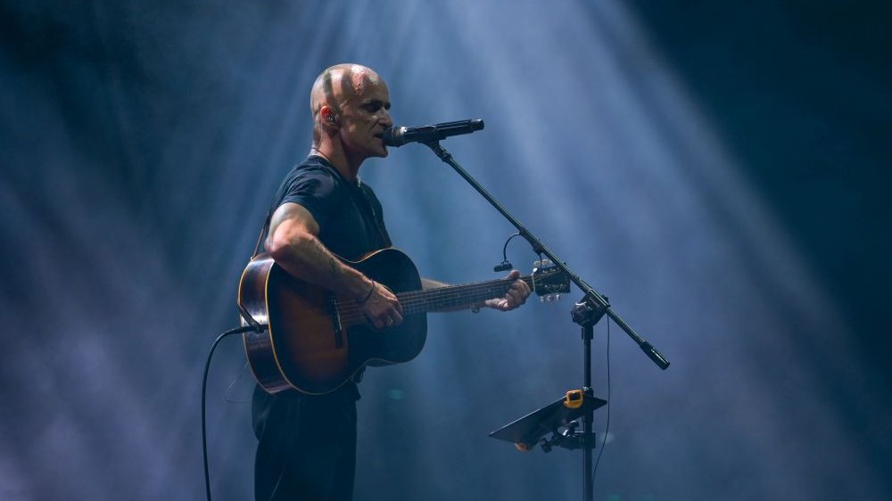 Artur Rojek stoi z gitarą przy statwywie, śpiewa do mikrofonu. Ma zamknięte oczy.