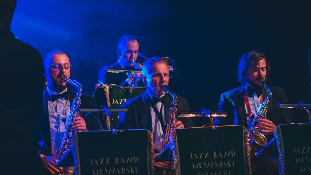 Orkiestra dęta, panowie grają na trąbkach i saksofonach. Na ich pulpitach widać materie, na których napisano złotymi literami Jazz Band Młynarski Masecki.
