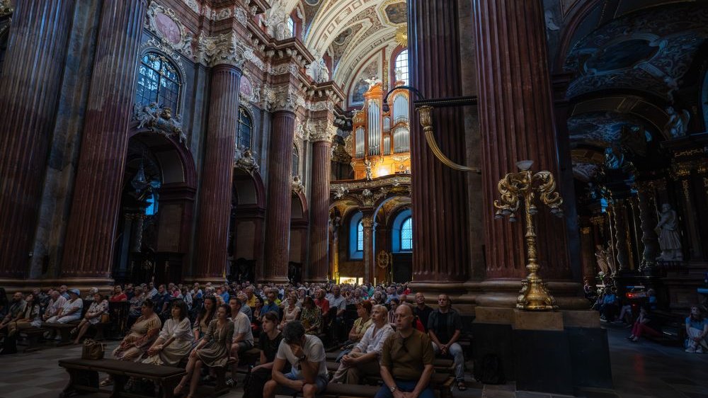 Słuchacze koncertu widziani z przodu fary, od strony ołtarza. Wydają się malutcy, obok nich widać wielkie świeczniki, o wiele większe kolumny i posągi w farze.