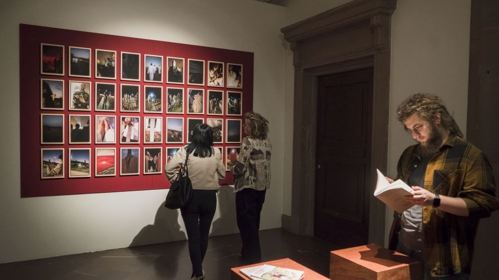 Tym razem fotografie na czerwonej ścianie oglądają dwie dziewczyny, za nimi stoi chłopak, przeglądający katalog.
