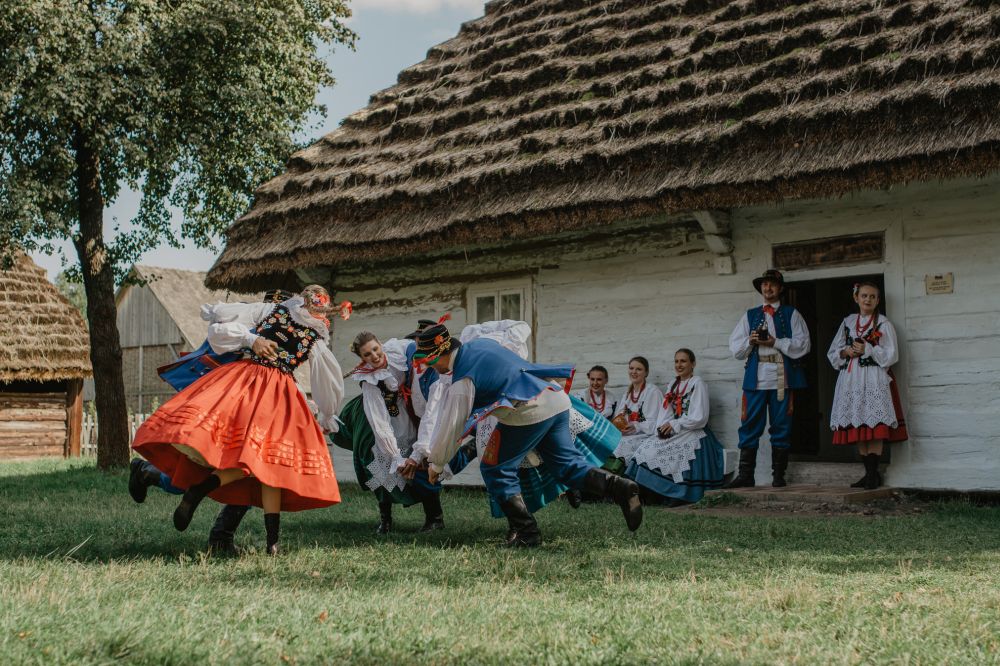 Tancerze w polskich strojach ludowych tańczą przed tradycyjną, krytą strzechą chatą chłopską. - grafika artykułu