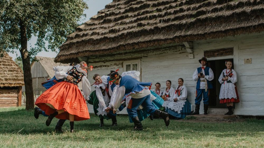 Tancerze w polskich strojach ludowych tańczą przed tradycyjną, krytą strzechą chatą chłopską.