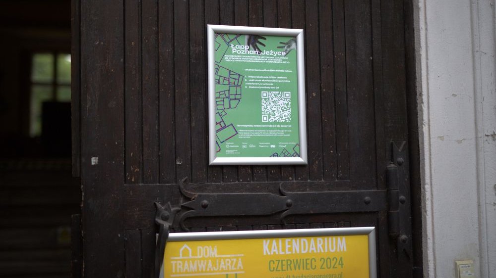 Zielony plakat z QR kodem, oprawiony w srebrną ramkę, wisi na drewnianym skrzydle drzwi.