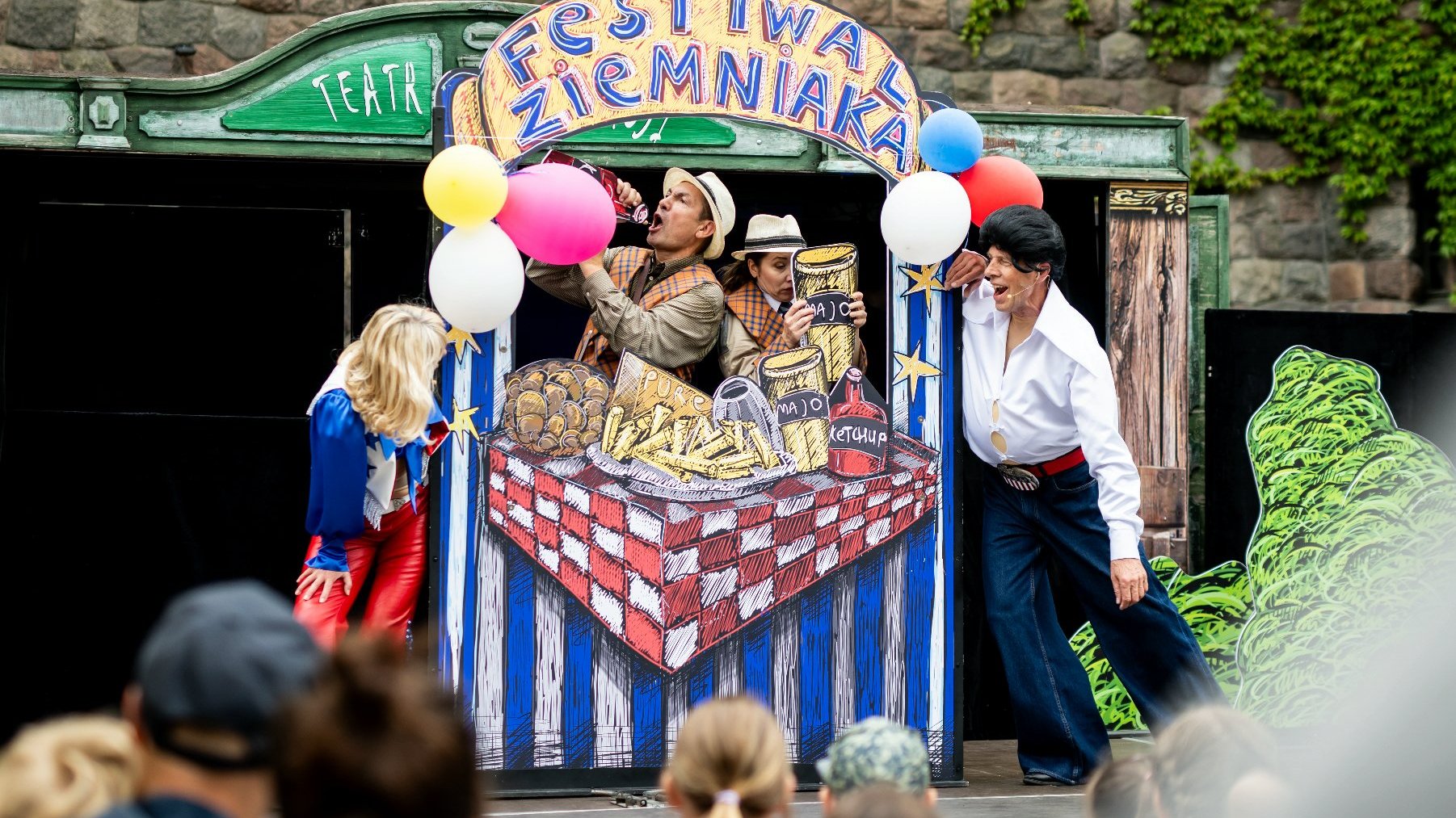 Scena ze spektaklu plenerowego na dziedzińcku Zamku. W centrum stragan z banerem Festiwalu Ziemniaka - płaski element scenografii. Dwójka zaaferowanych aktorów/detektywów udaje sprzedawców. Dwoje innych jest na drugim planie, podtrzymuje scenogarfię/stragan.