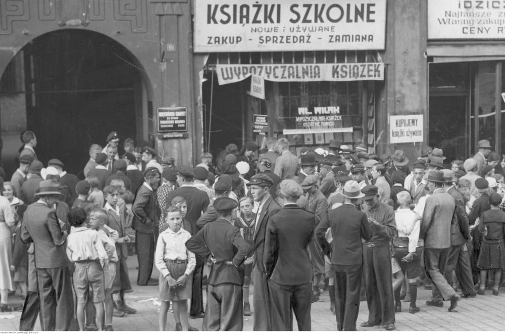 Czarno-białe zdjęcie tłumu dorosłych i dzieci stojących na ulicy przed komisem książek używanych. Na budynku sklepu wisi dużo reklamowych szyldów. - grafika artykułu