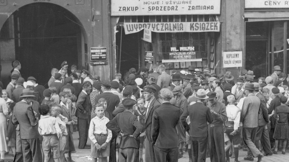 Czarno-białe zdjęcie tłumu dorosłych i dzieci stojących na ulicy przed komisem książek używanych. Na budynku sklepu wisi dużo reklamowych szyldów.