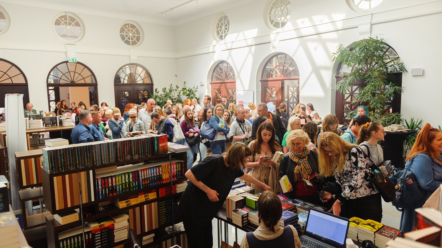 Tłum ludzi w sali, gdzie jest dużo książek. Oglądają i kupują książki.