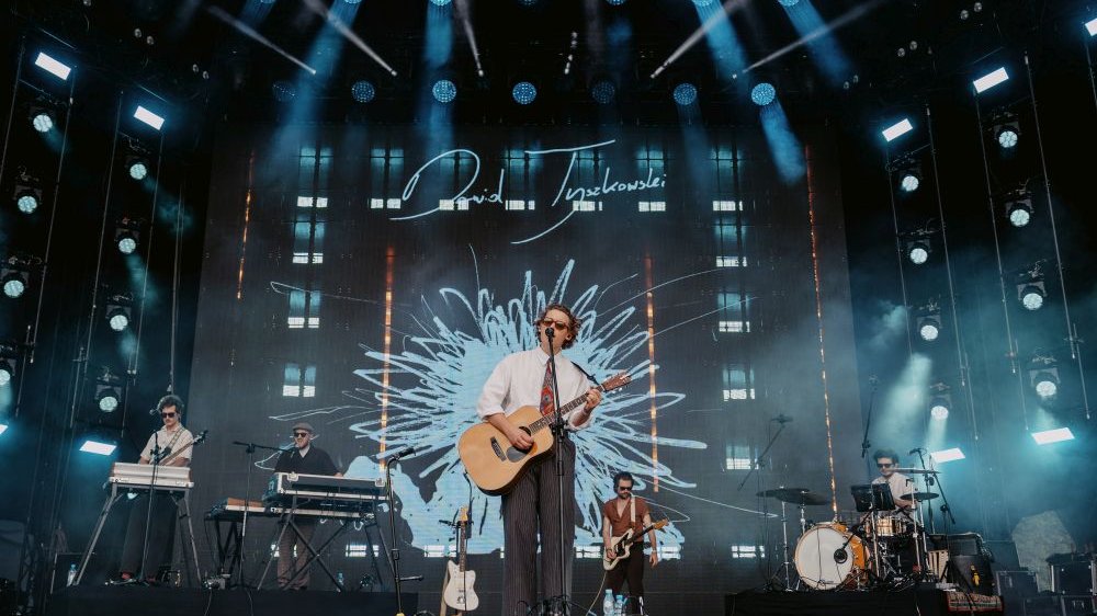 Artysta ma na sobie ciemne okulary, białą koszulę i kolorowy krawat oraz garniturowe spodnie. Śpiewa, grając na gitarze, za nim widać resztę zespołu - każdy muzyk ma na twarzy czarne okulary a na głowie podobną fryzurę z podkręconymi włosami.