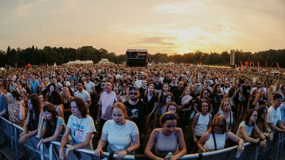 Publiczność, przeważają w niej młode kobiety, ale są też mężczyźni i chłopcy. Za publicznością w plenerze widać parkowe drzewa oraz zachodzące za nimi słońce.