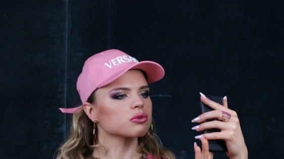 Ekran podpisany jako "Type 8. Sex Doll". Widnieje na nim kobieta w różowym dresie i czapce z napisem "Versace", która robi sobie zdjęcie telefonem komórkowym. Ma długie paznokcie, złotą biżuterię i mocny makijaż na twarzy. Tło za nią jest czarne.