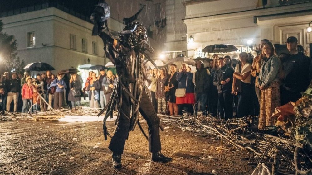 Na środku placu stoi postać ubrana na czarno, widzowie patrzą tylko na nią. Podczas spektaklu padał deszcz - chodnik jest mokry i powstają na nim kałuże, a wielu widzów ma nad sobą parasole.