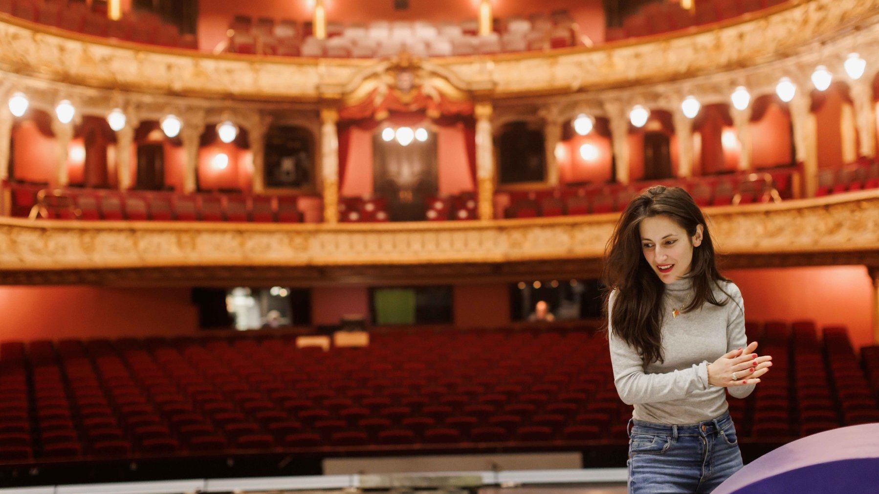 Kobieta z długimi, rozwianymi włosami prowadzi próbę w teatrze, za nią puste krzesła widowni