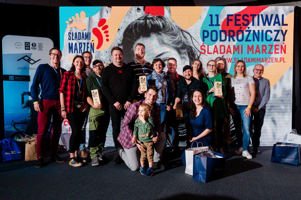 Grupa uśmiechniętych ludzi pozuje przy plakacie reklamującym "Śladami marzeń", niektórzy trzymają w rękach statuetki. - grafika artykułu