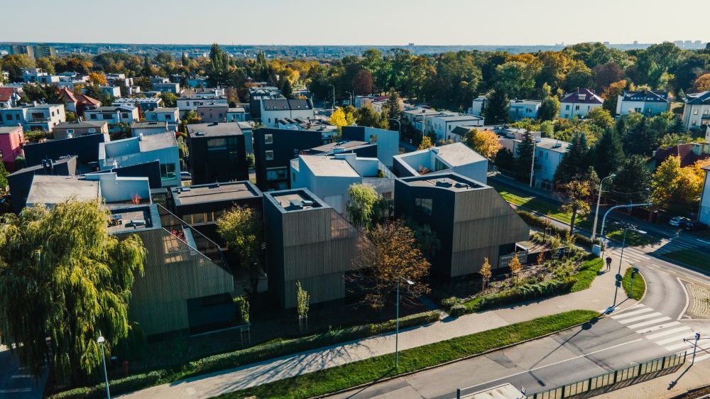 Małe osiedle budynków wykończonych drewnem wpisane w architekturę ulicy w okolicy Cytadeli. Dookoła dużo drzew i zieleni.