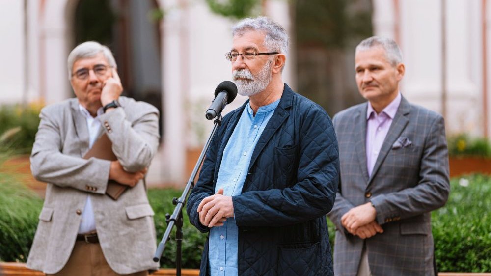 Siwy mężczyzna z brodą przemawia do mikrofonu na świeżym powietrzu. Za nim stoją dwaj przedstawiciele miasta.