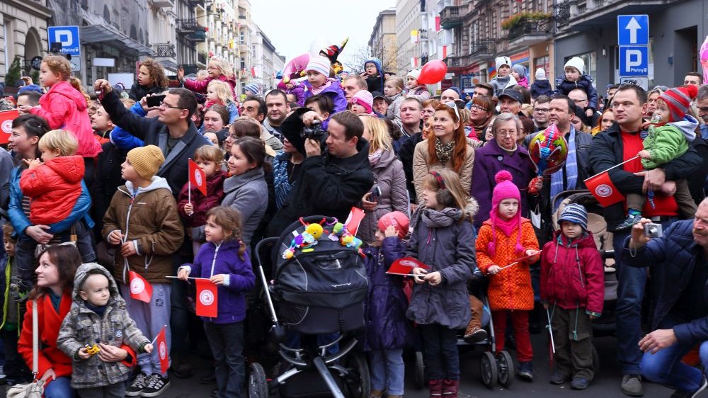 Tłum ludzi ogląda świąteczny pochód. Jest dużo dzieci, mają w rękach chorągiewki i baloniki.