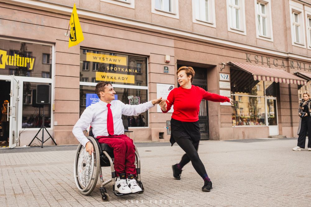 Kobieta w czerwonym sweterku i mężczyzna w białej koszuli i z czerwonym krawatem tańczą na ulicy. Mężczyzna porusza się na wózku inwalidzkim. - grafika artykułu