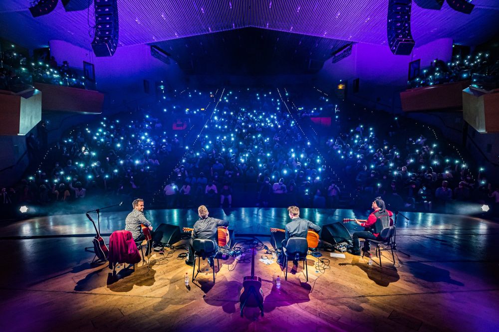 Czterech członków zespołu w trakcie koncertu gra na gitarach na scenie. Przed nimi publiczność rozświetlona latarkami w telefonach komórkowych. - grafika artykułu