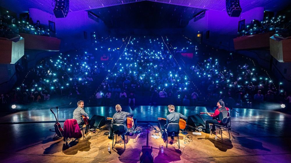 Czterech członków zespołu w trakcie koncertu gra na gitarach na scenie. Przed nimi publiczność rozświetlona latarkami w telefonach komórkowych.