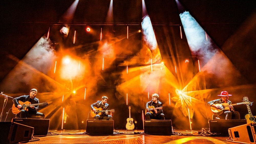 Czterech gitarzystów występuje na scenie. Z góry pada na nich pomarańczowe światło.