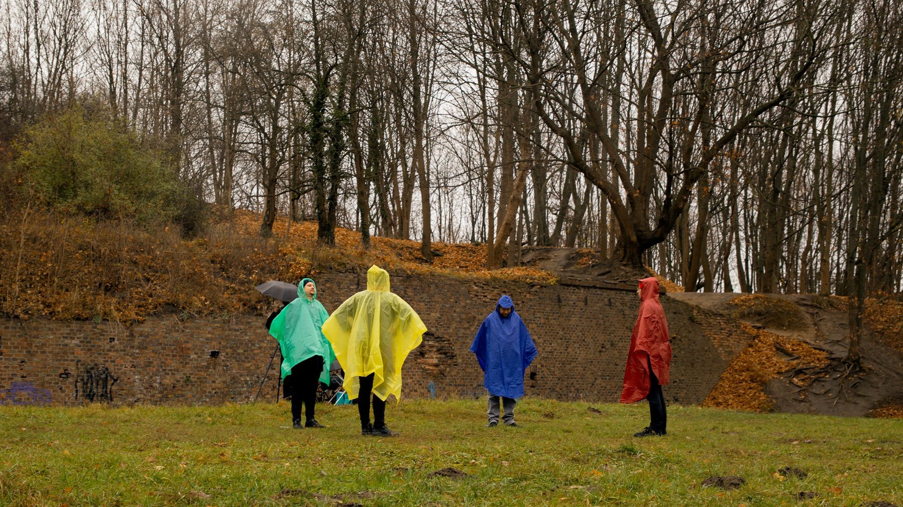 Czwóka mężczyzn stoi w okręgu. Mają kolejno: żółtą, czerwona, niebieska i zieloną peleryną przeciwdeszczową. Stoją na trawie, za nimi stary mur i drzewa.