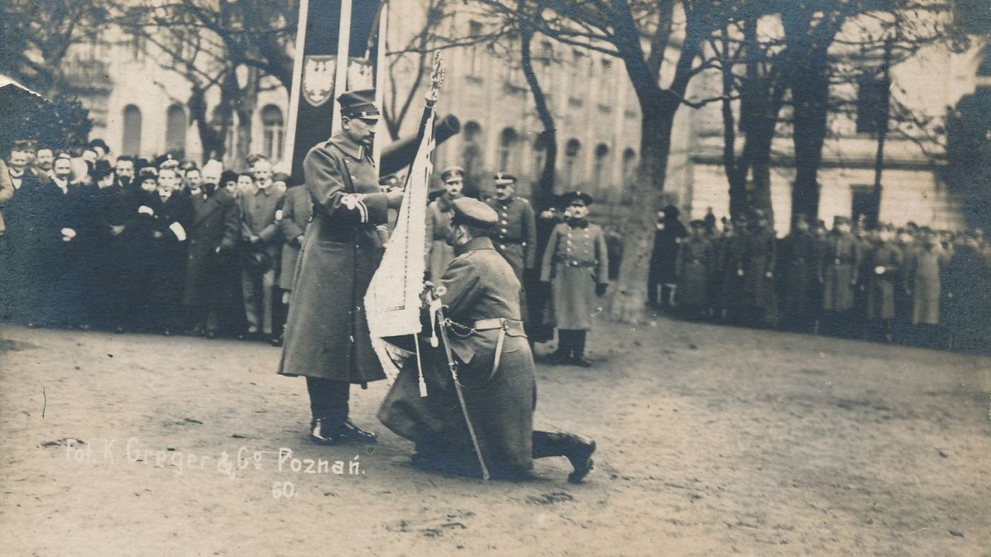 Żołnierz klęcząc przyjmuje sztandar z rąk dowódcy