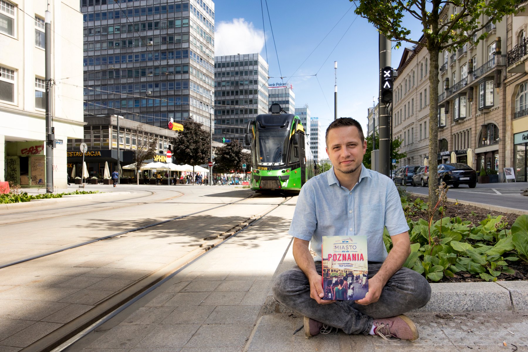 Młody, uśmiechnięty mężczyzna siedzi na krawężniku z książką na kolanach - grafika artykułu