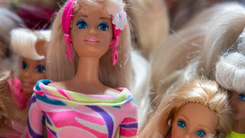 Lalka barbie w różowej opasce i kolorowej bluzce obok innych Barbie. Wszystkie mają blond włosy.