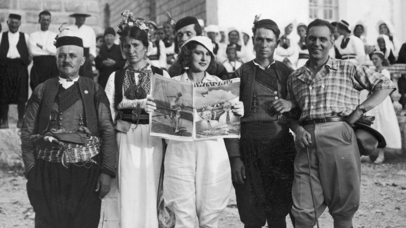 Scena z filmu. Rząd ludzi w różnym wieku. Młoda kobieta w środku czyta magazyn i się uśmeicha. Wśród nich jest para ludzi w stroju ludowym.