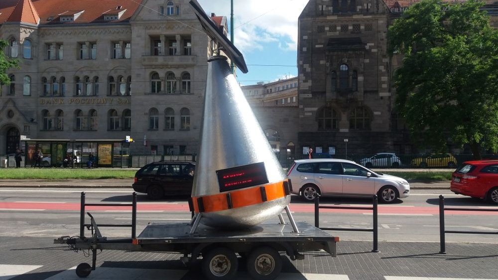 Srebrna boja, przypominająca nieco statek kosmiczny o stożkowym kształcie, stoi na samochodowej przyczepce przed CK Zamek przy ulicy Święty Marcin.