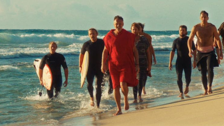 Grupa mężczyzn z deskami do surfowania idzie brzegiem plaży. Za nimi wzburzone fale.